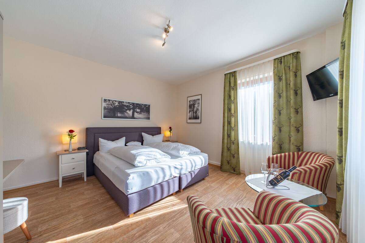 A hotel room with a bed, bedside tables, a TV, a dining area with a table and chairs, and a window with curtains.