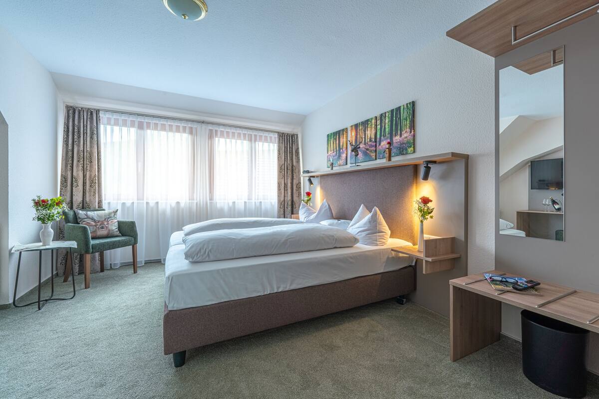 A modern hotel room with a large bed, bedside lamps, a desk, a chair, and a large window with curtains.