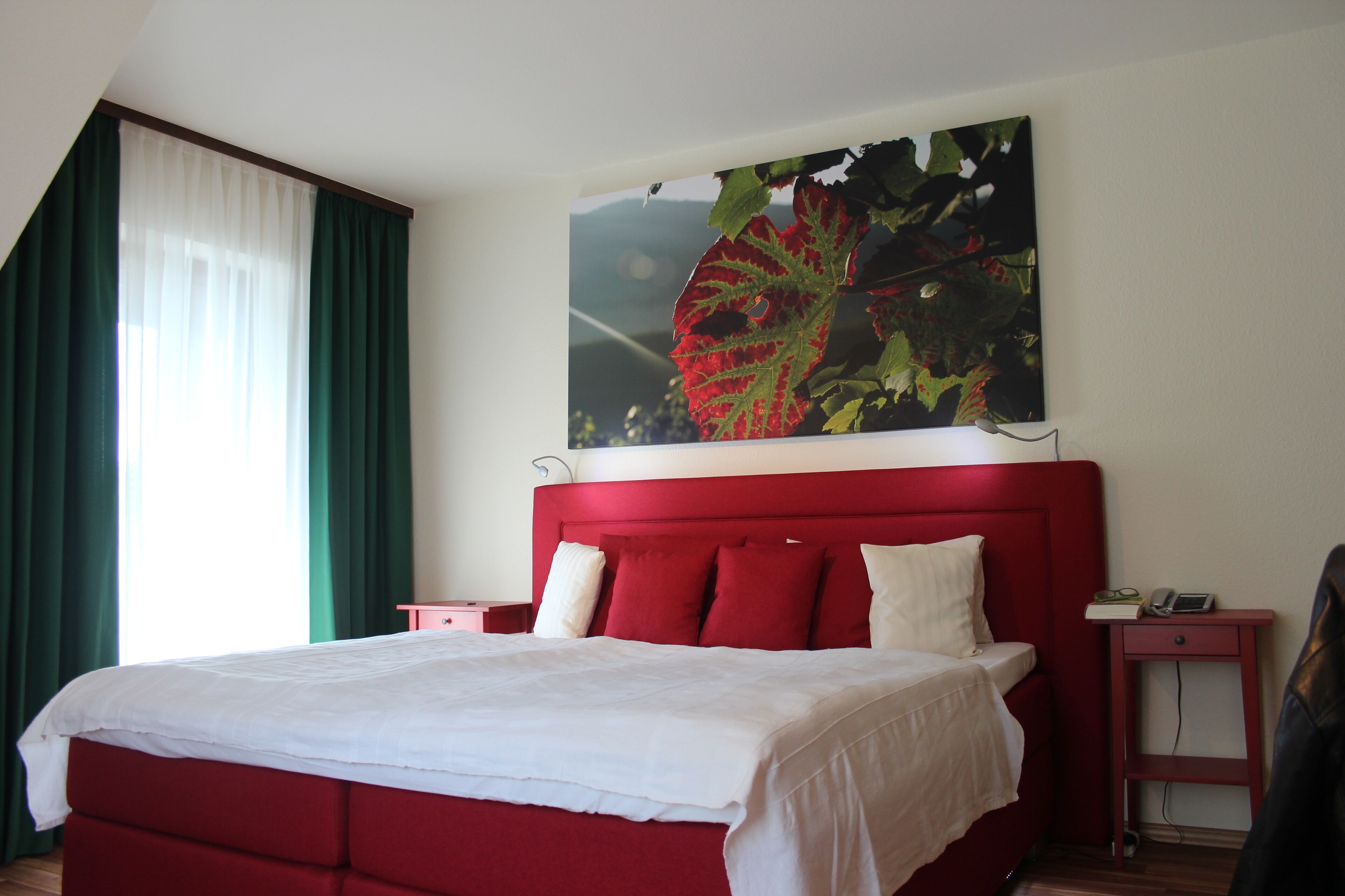 A red bed with white linens, a nightstand with a phone and book, and a painting of a leaf on the wall.