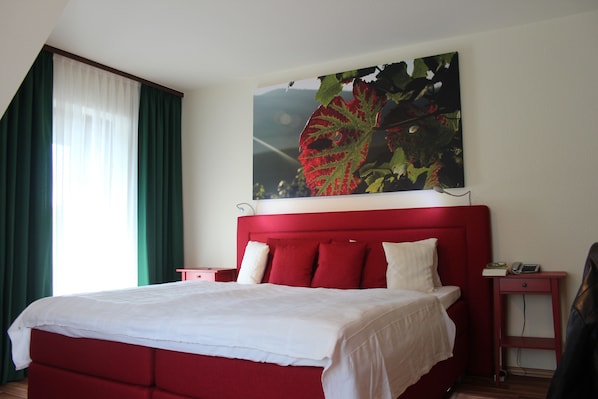 Cama roja con ropa de cama blanca, mesita de noche con teléfono y libro, y un cuadro de una hoja en la pared.