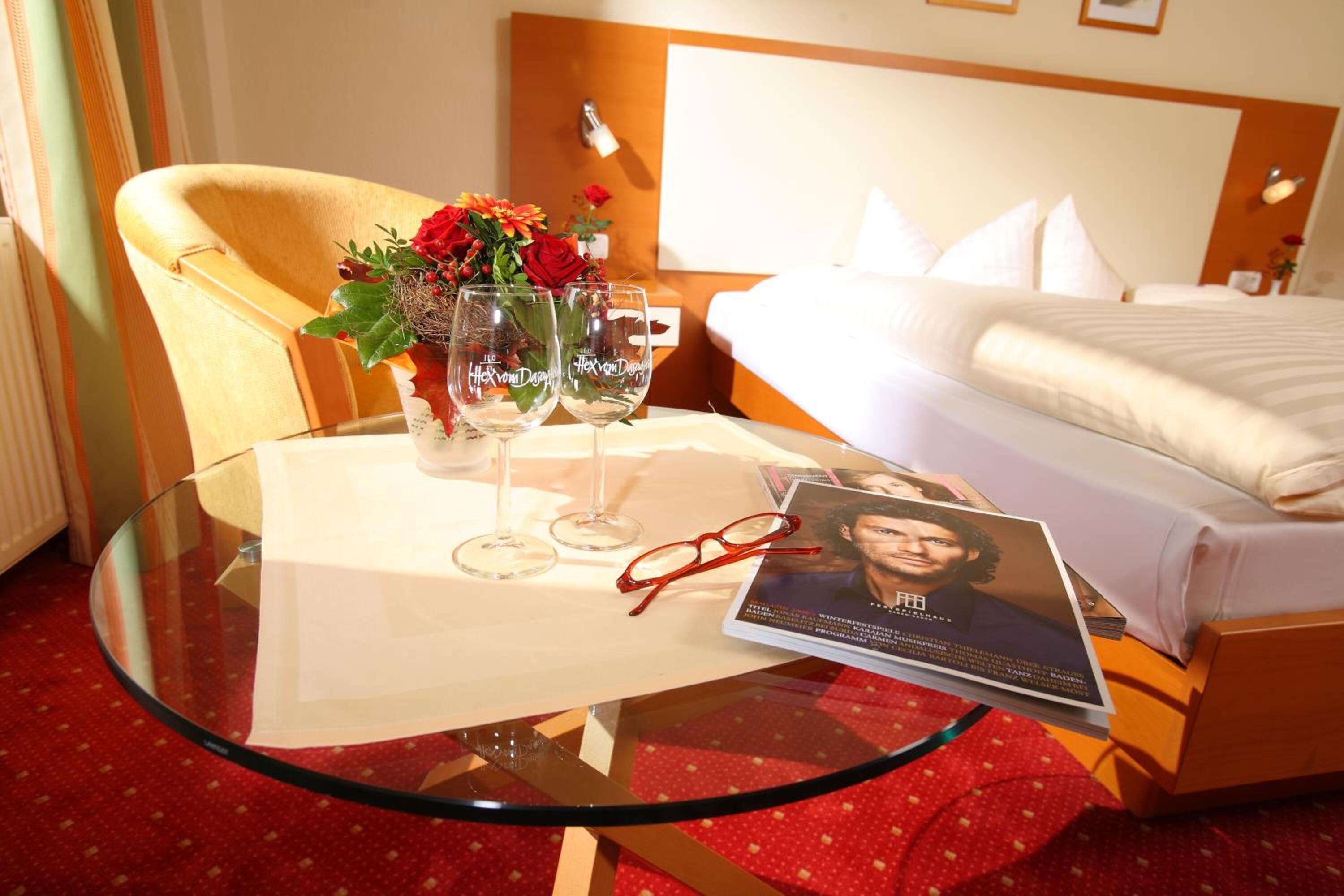 A hotel room with a bed, a glass table with a brochure, two wine glasses, and a vase with flowers.