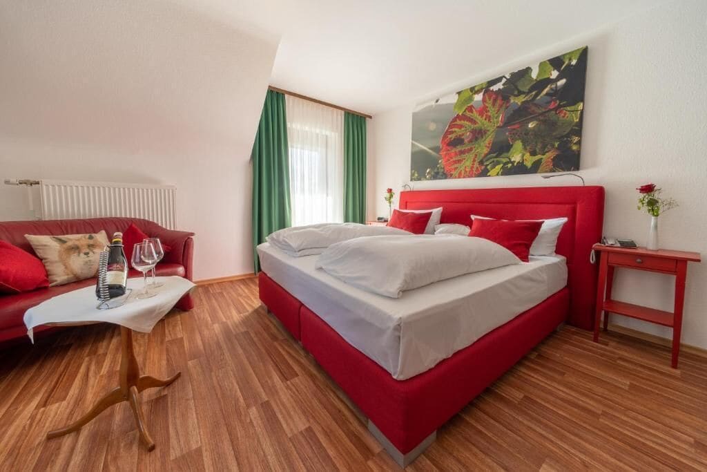 Un cuarto de hotel con una cama roja, ropa de cama blanca, un sofá rojo, una mesita con una botella y un vaso, y un cuadro en la pared.
