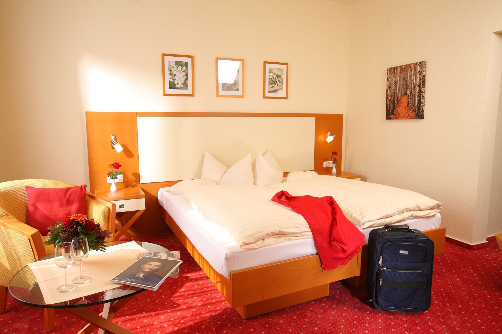 Ein Hotelzimmer mit Bett, Nachttischen, einem Sessel, einem Glastisch mit roter Tischdecke und einem Koffer.