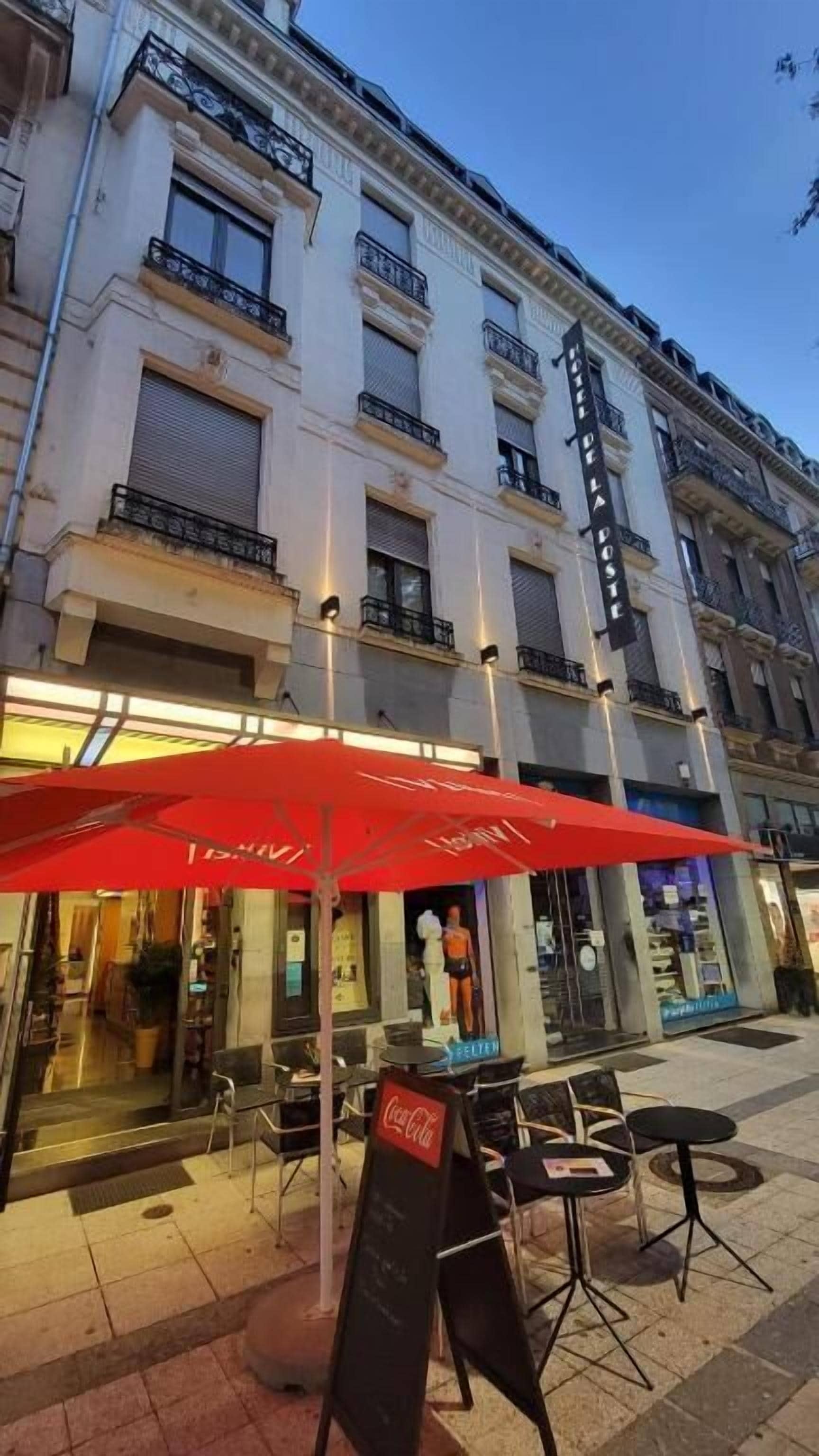 Un edificio de varios pisos, con mesas al aire libre y sombrillas rojas, y un letrero de Coca-Cola.