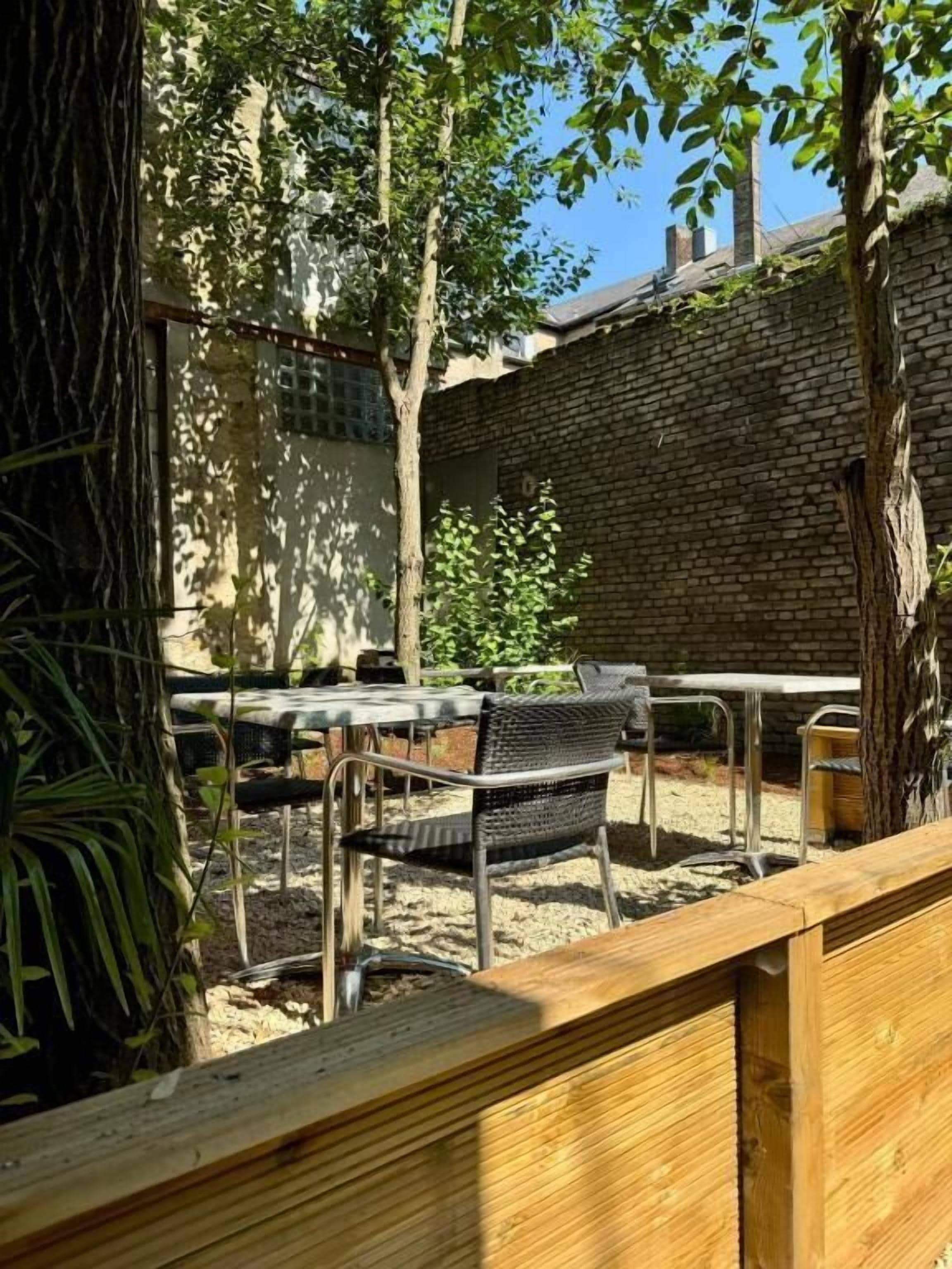 Un área de estar al aire libre con sillas de metal y una mesa de madera, rodeada de árboles y un muro de ladrillo.