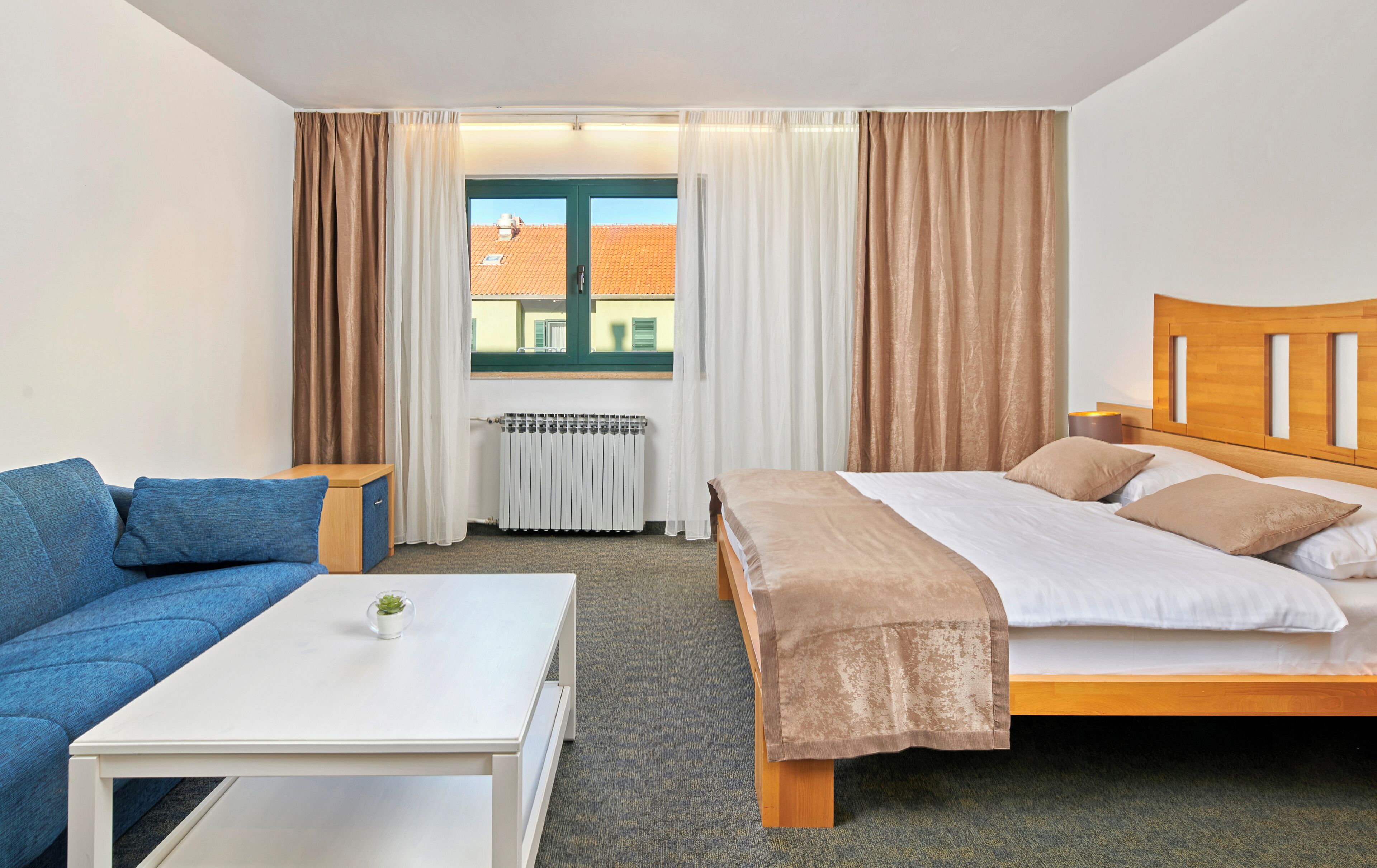 A hotel room with a bed, a sofa, a small table, a radiator, and a window with curtains.