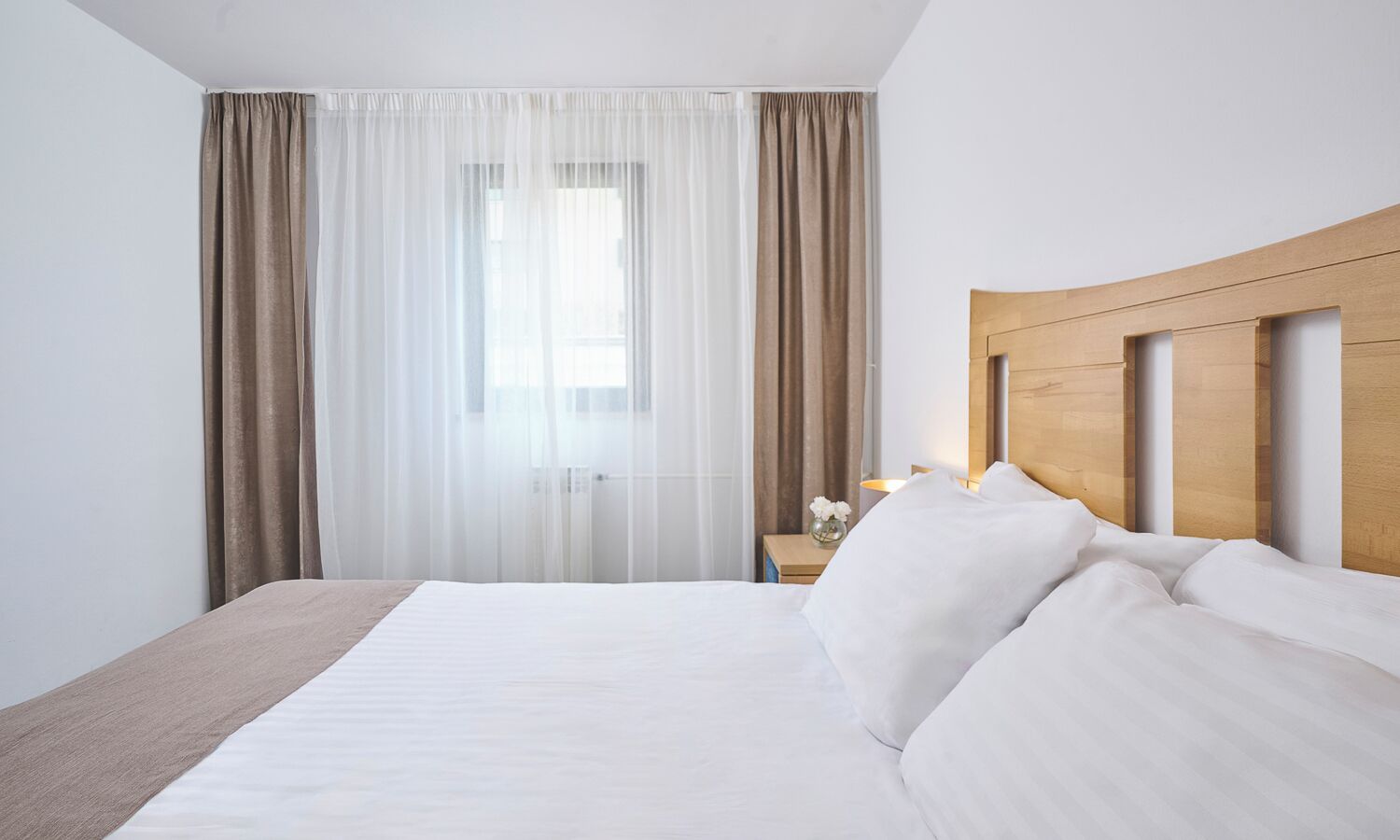 A hotel room with a large bed, a wooden headboard, a nightstand with a lamp, and sheer curtains.