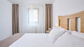 A hotel room with a large bed, a wooden headboard, a nightstand with a lamp, and sheer curtains.