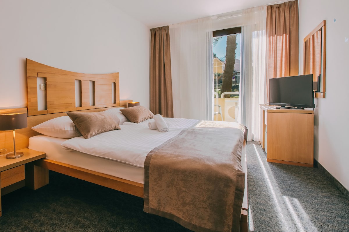 Quarto de hotel com cabeceira de madeira, cama com roupa de cama branca, luminária de cabeceira, televisão sobre suporte de madeira e janela com cortinas.