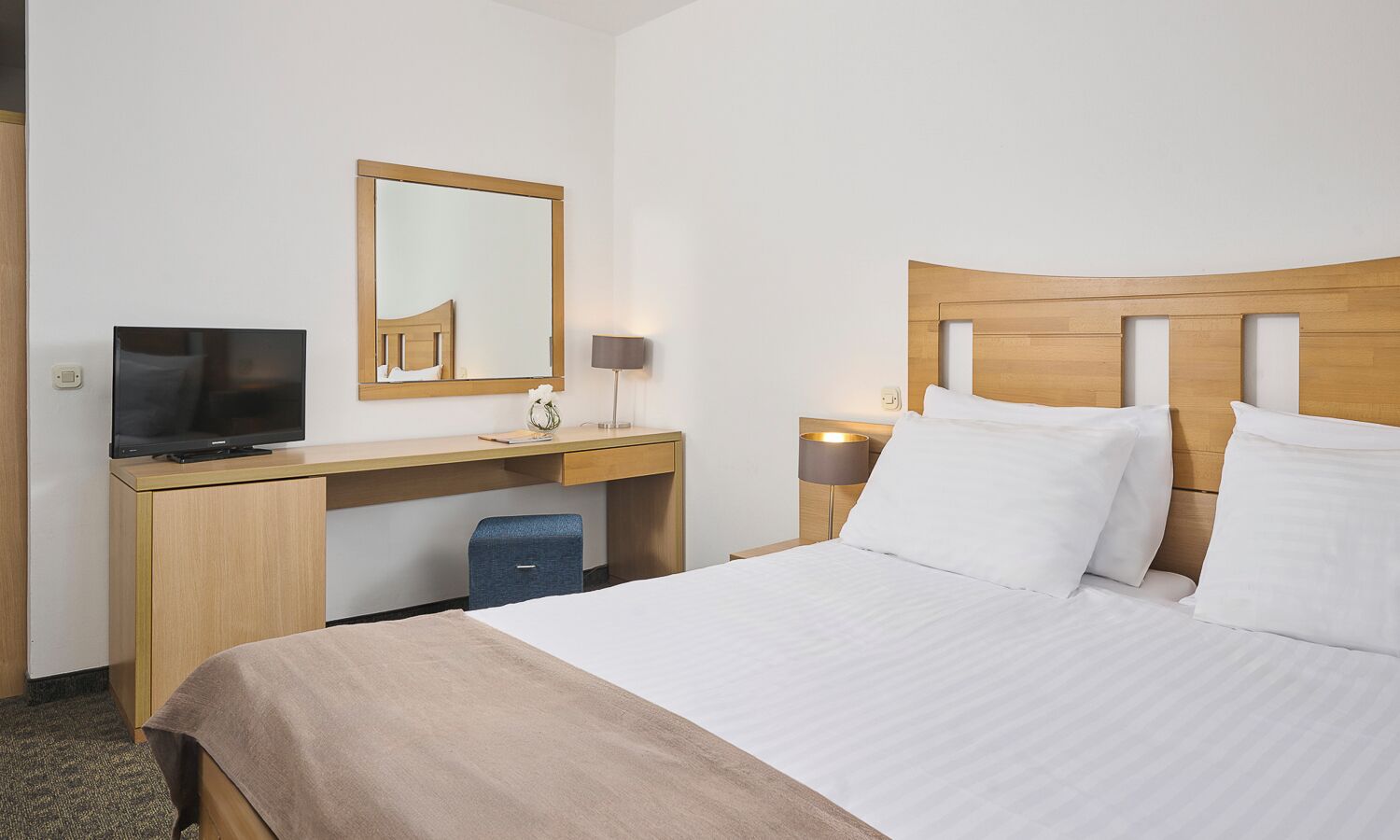 A hotel room with a bed, a desk with a TV, a mirror, and bedside lamps.