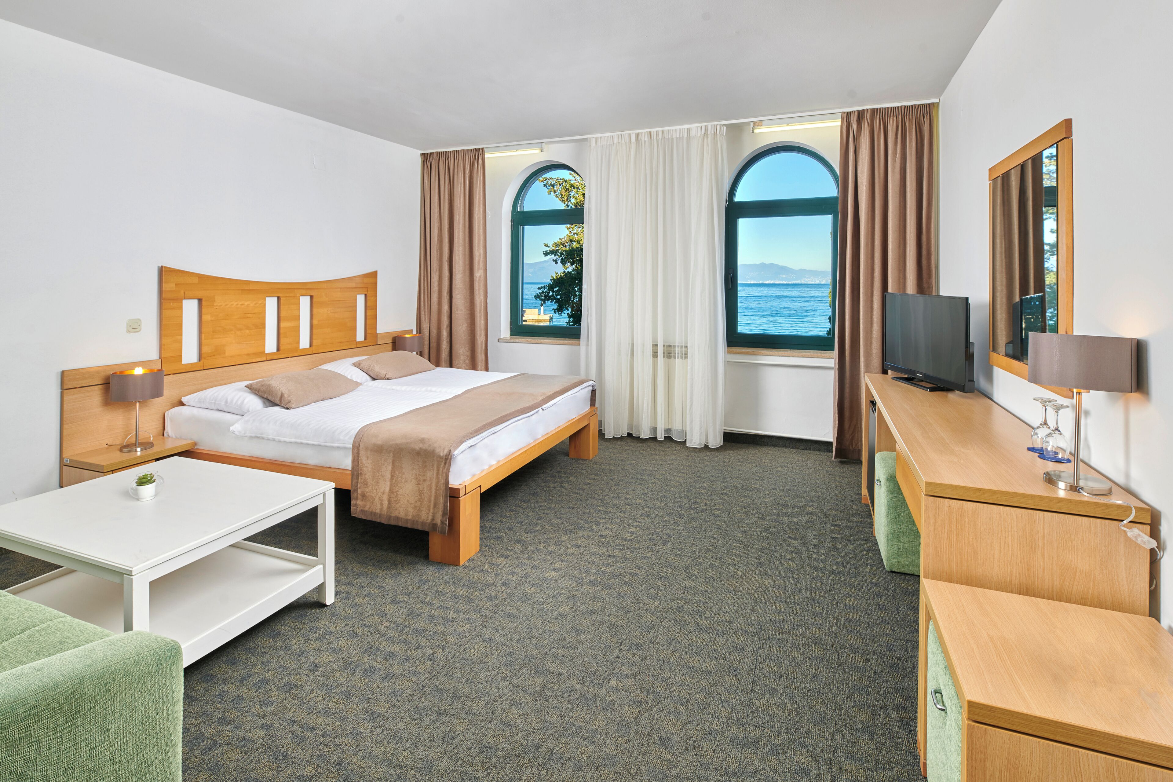 A hotel room with a bed, a desk, a TV, and large windows with curtains.