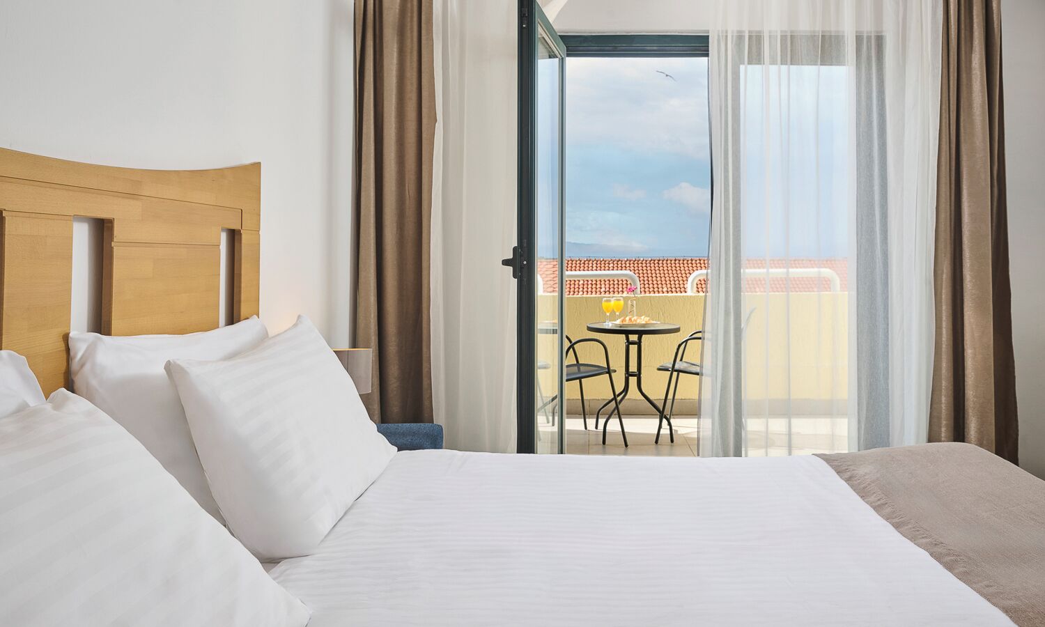 A hotel room with a bed, a view of the terrace, and a table with chairs outside.