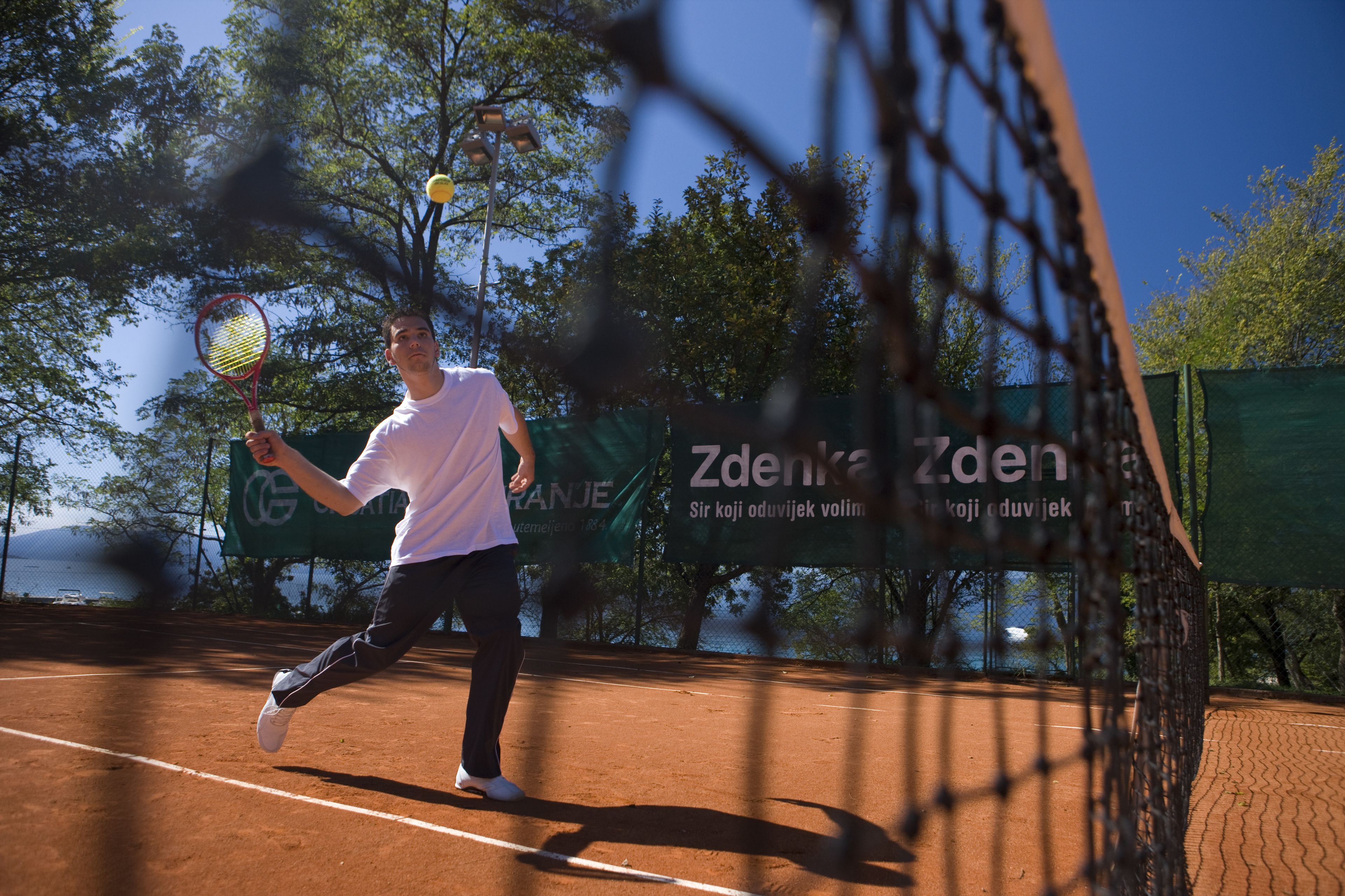 A tennis court with a player in mid-action, a net, and a banner with text in the background.