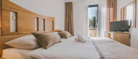 A hotel room with a wooden headboard, a bed with white and beige bedding, a flat-screen TV on a wooden stand, and a balcony with curtains.