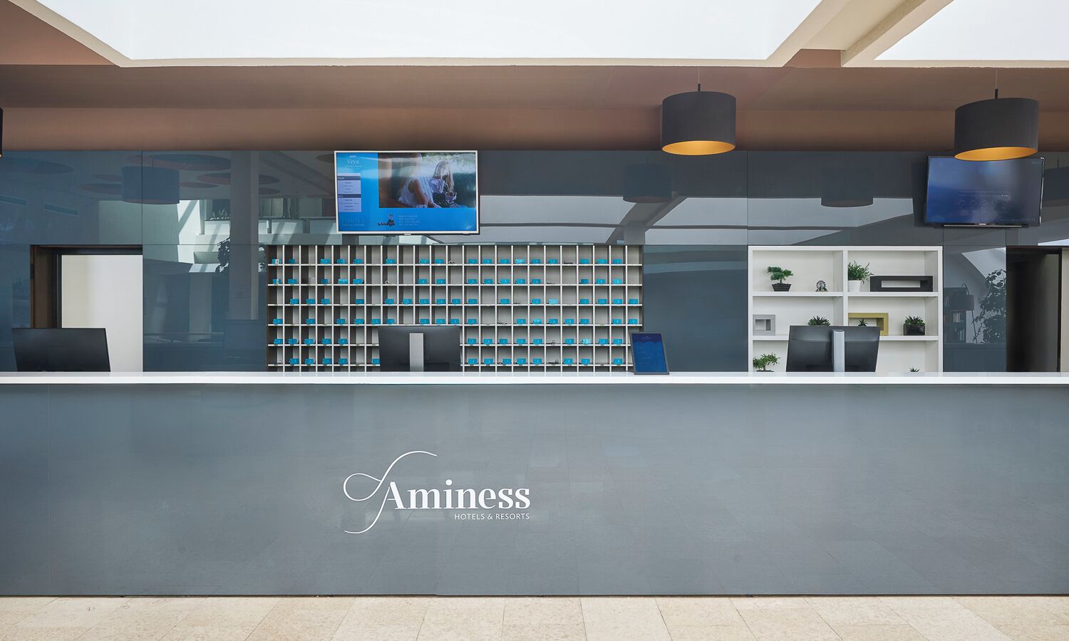 Ein Empfangsschalter mit einem digitalen Bildschirm, Regale mit blauen Gegenständen und ein Logo von Aminess Hotels & Resorts.