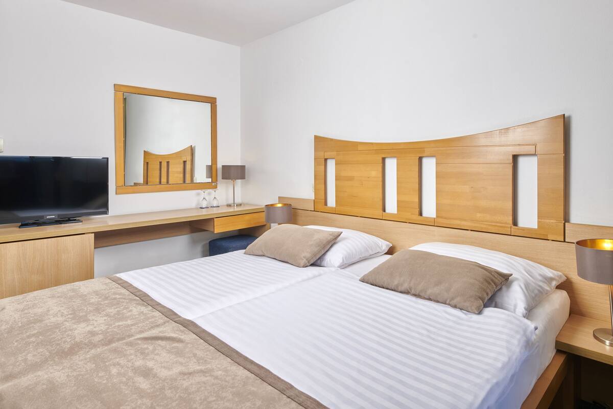 Quarto de hotel com cama, escrivaninha com televisão, espelho e mesa de cabeceira com abajur.