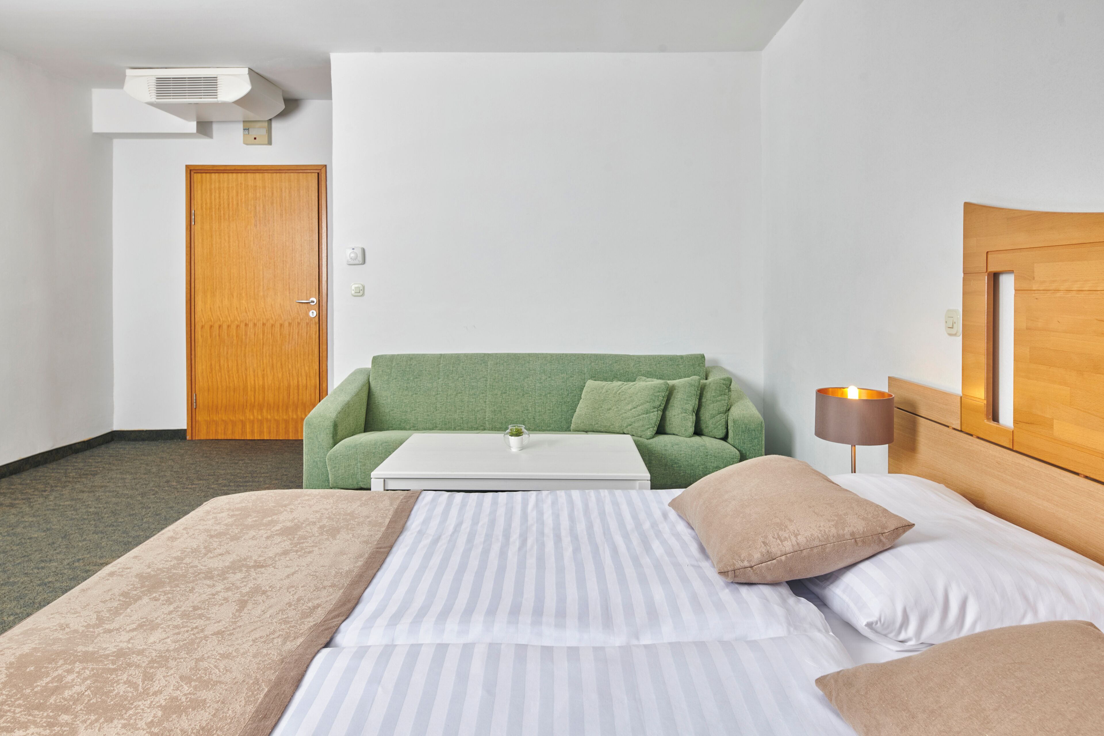 Een hotelkamer met een bed, een groene bank, een klein tafeltje, een lamp en een houten deur.