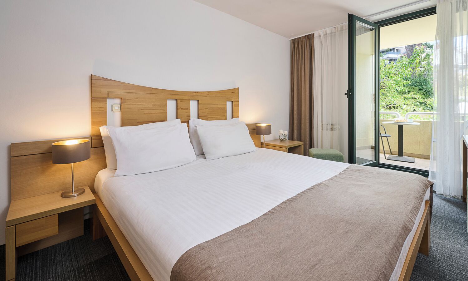Habitación de hotel con cama, dos mesitas de noche, lámpara, ventana con cortinas y vista a zonas verdes.
