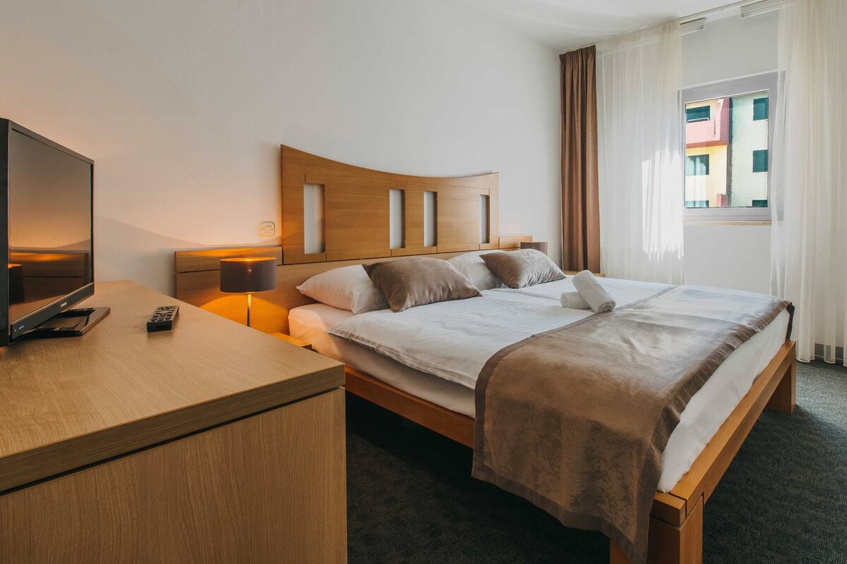 Quarto de hotel com cama de madeira, televisão sobre uma mesinha de madeira e janela com cortinas.