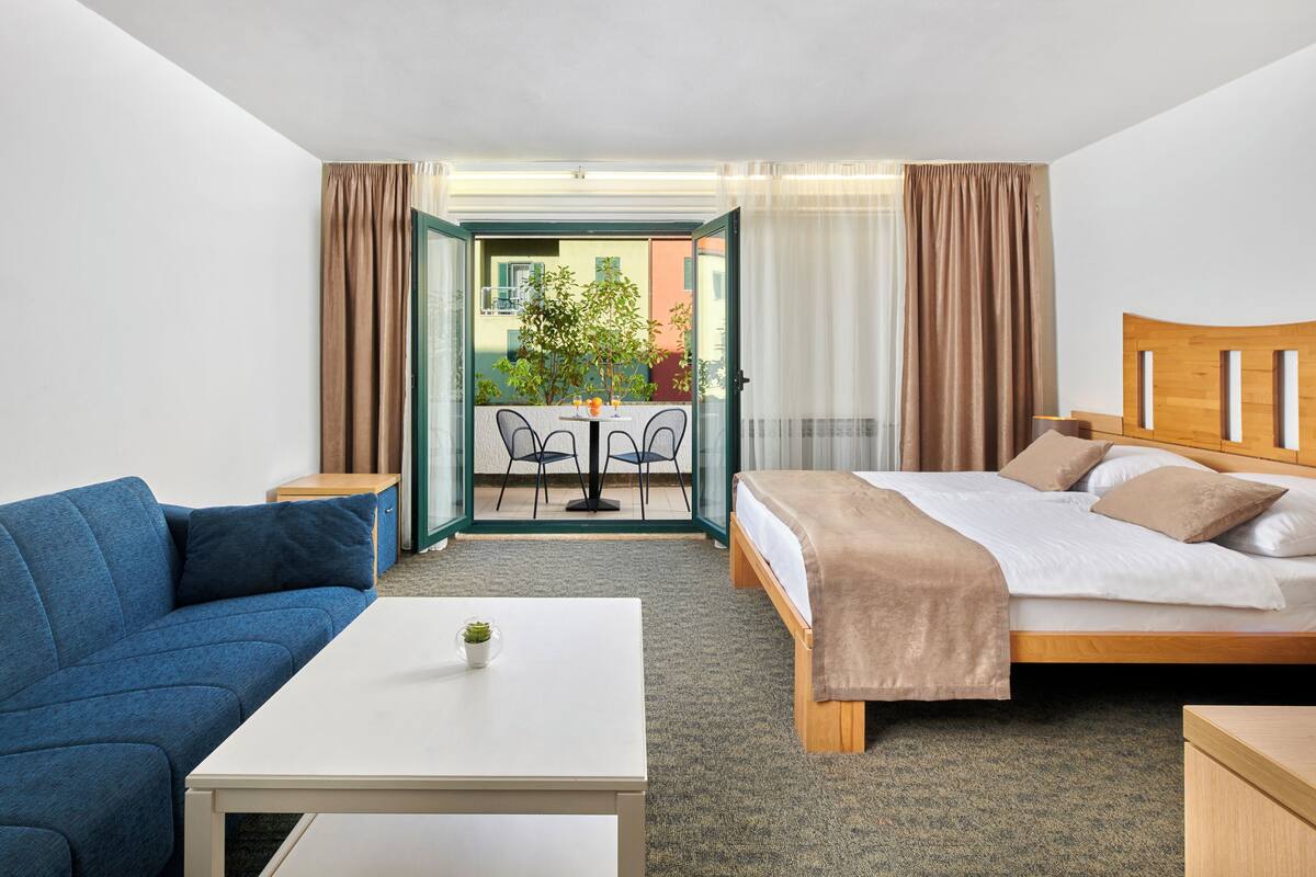 Quarto de hotel com uma cama, um sofá azul, uma mesa pequena com uma planta e vista para a varanda externa.