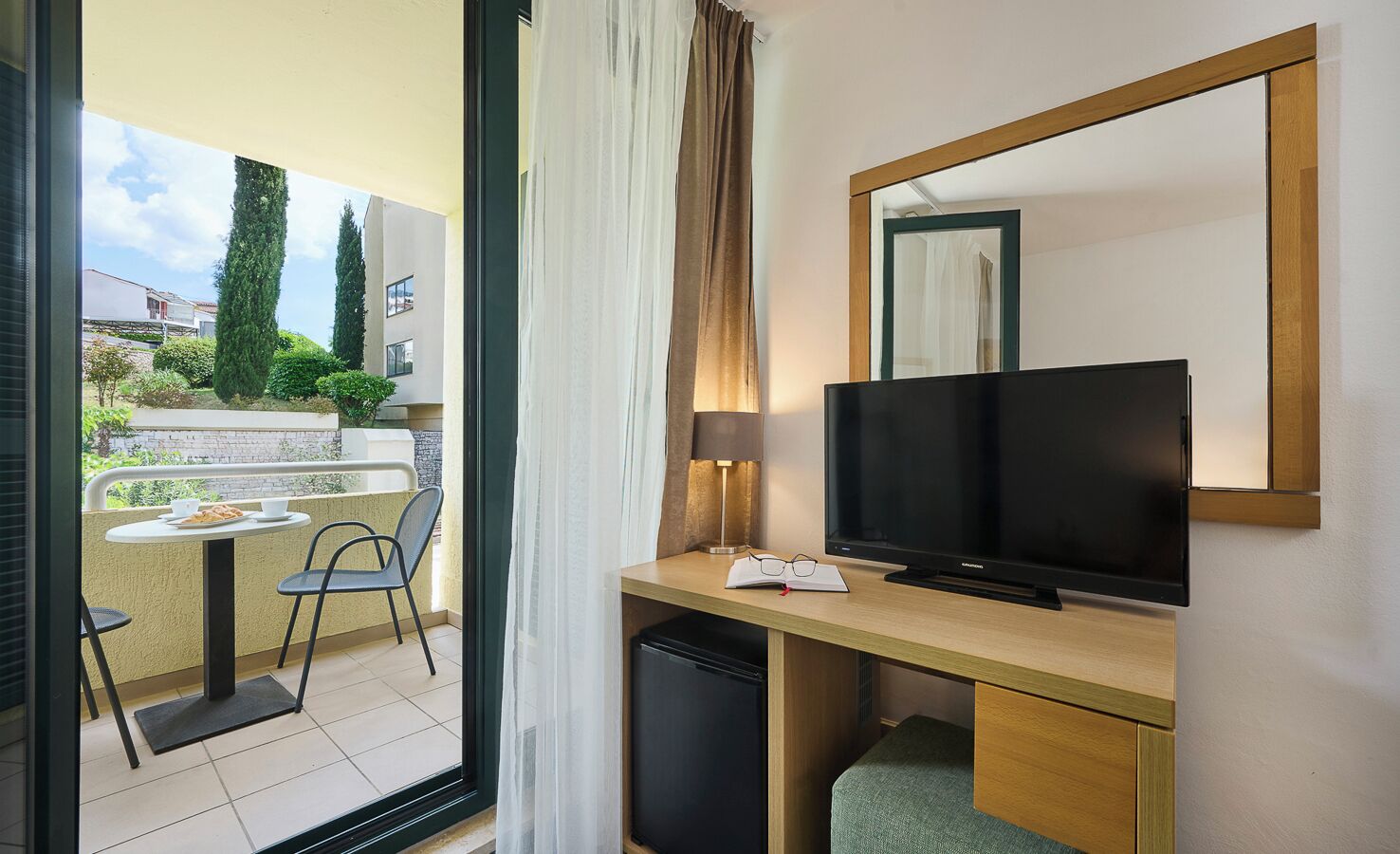 Une chambre d’hôtel équipée d’une télévision, d’un bureau, d’une chaise, d’une petite table avec une tasse et une soucoupe, et offrant une vue sur le balcon extérieur.