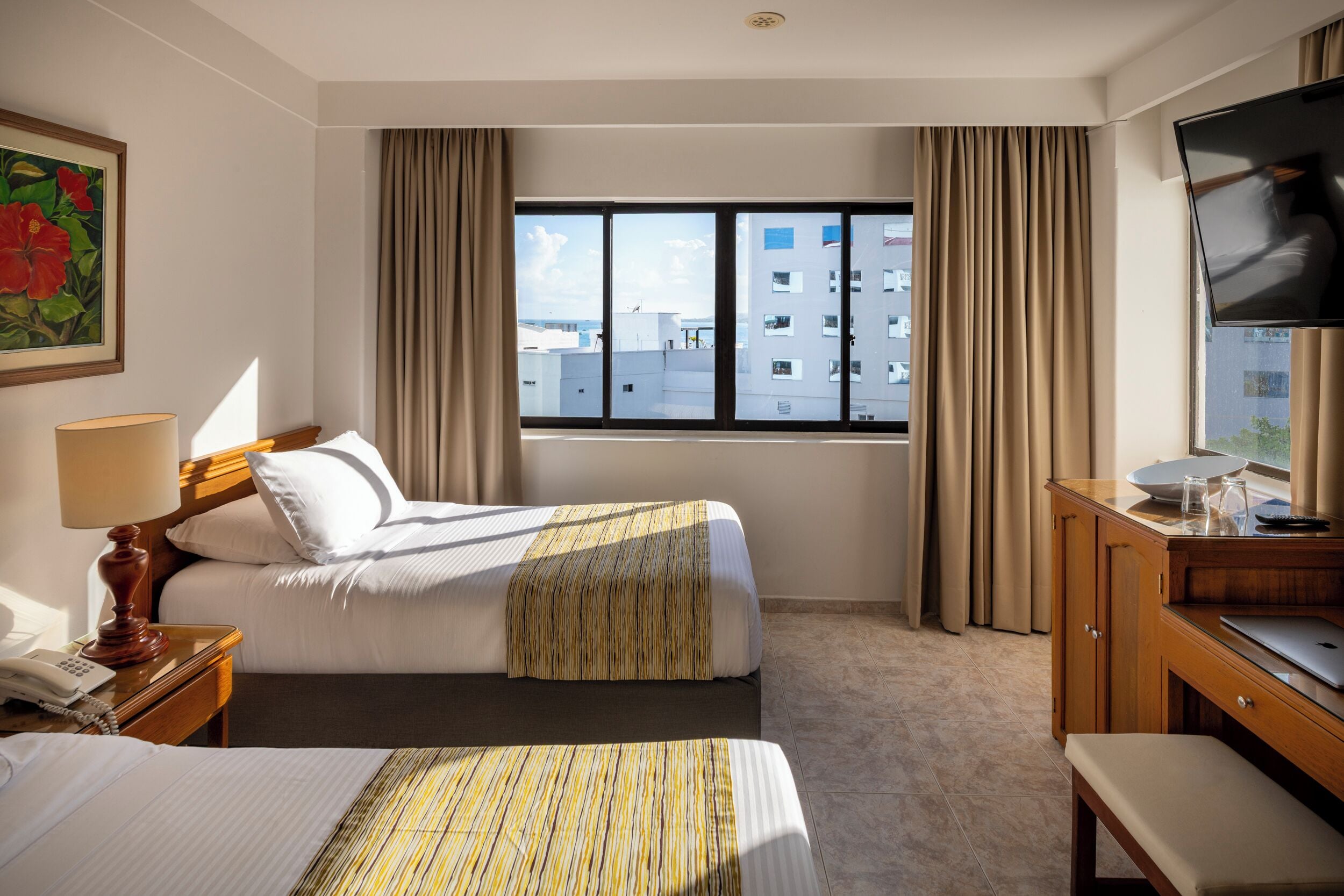 A hotel room with two beds, a desk with a TV, a window with curtains, and a painting on the wall.