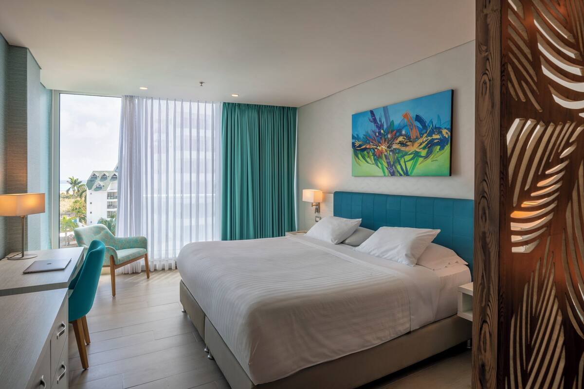 A hotel room with a large bed, a desk with a chair, a painting on the wall, and a view of the city.