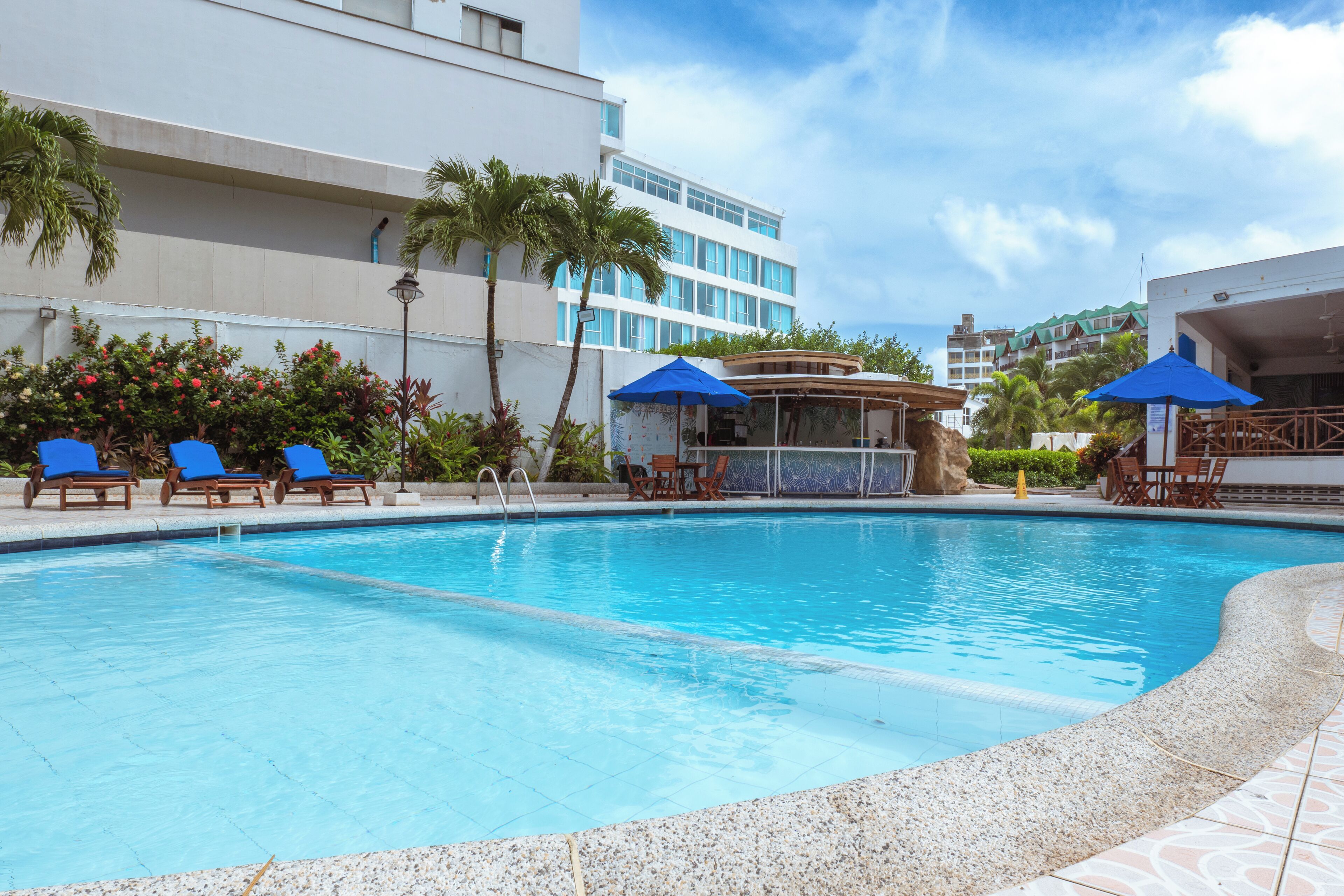 A swimming pool with lounge chairs and umbrellas, surrounded by buildings and palm trees.