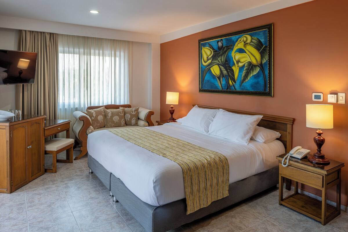 A hotel room with a large bed, a desk with a chair, a television, and a painting on the wall.