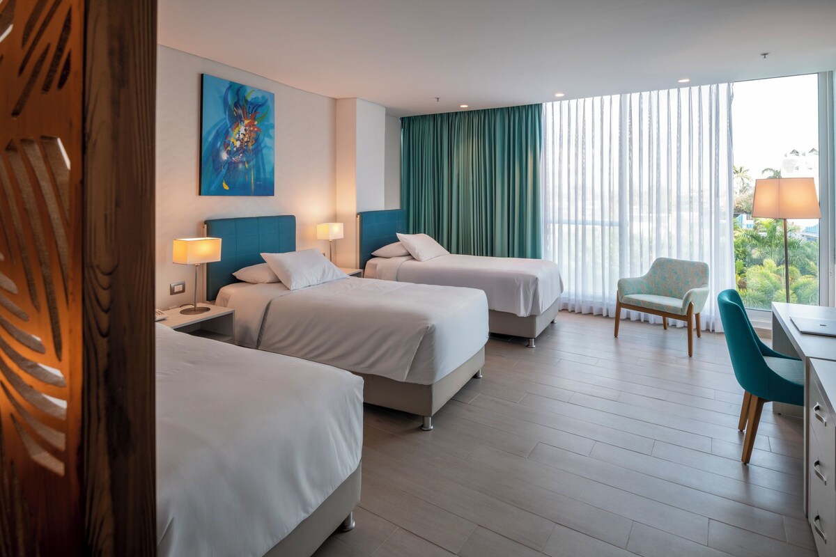 A hotel room with two beds, a desk, a chair, and a large window with curtains.