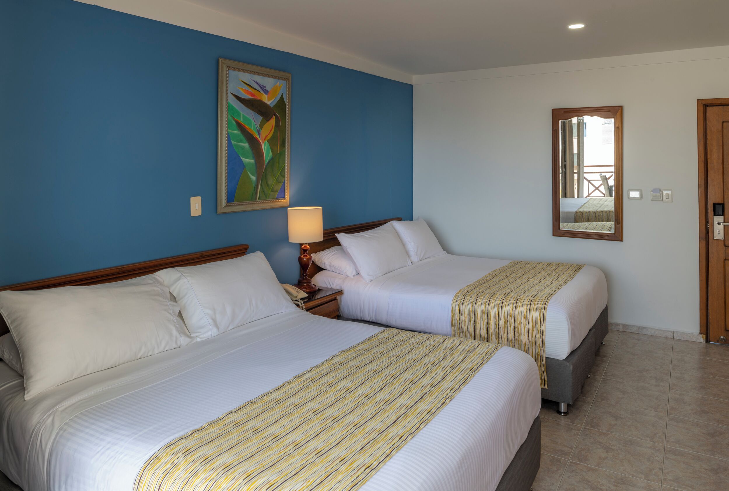 Habitación de hotel con dos camas, una mesita de noche con lámpara, un cuadro en la pared y un espejo.