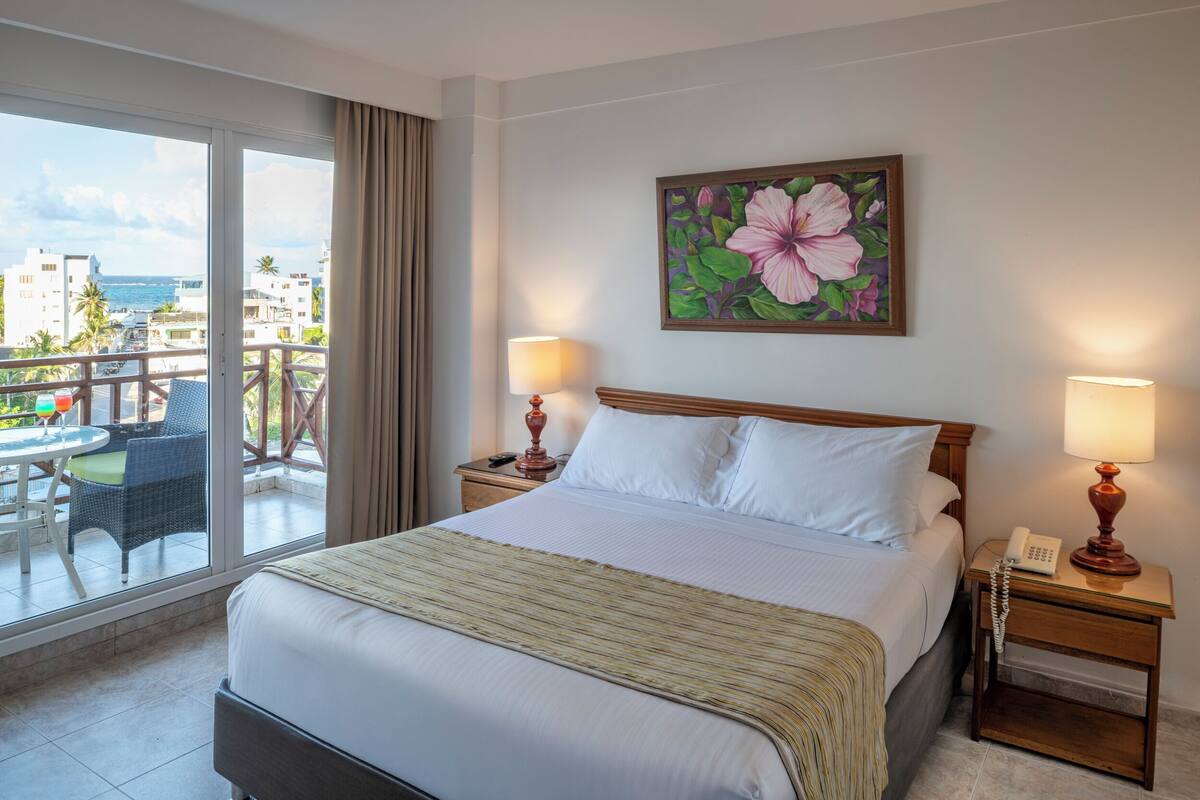 A hotel room with a bed, two bedside lamps, a painting of flowers, a balcony with a table and chairs, and a view of the sea and buildings.