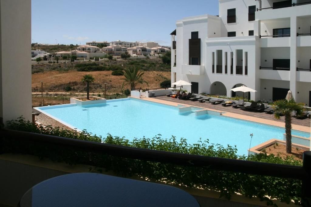 A modern hotel with a pool, sun loungers, and a view of the surrounding landscape.