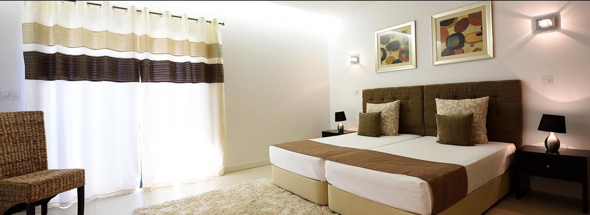 Quarto com cama de casal, duas mesas de cabeceira, uma cadeira e uma janela grande com cortinas.