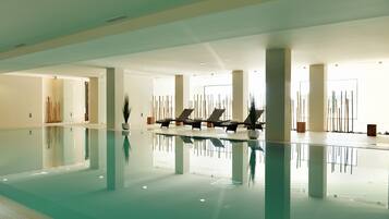 Una amplia piscina interior con agua cristalina, rodeada de sillas de descanso y plantas en macetas.