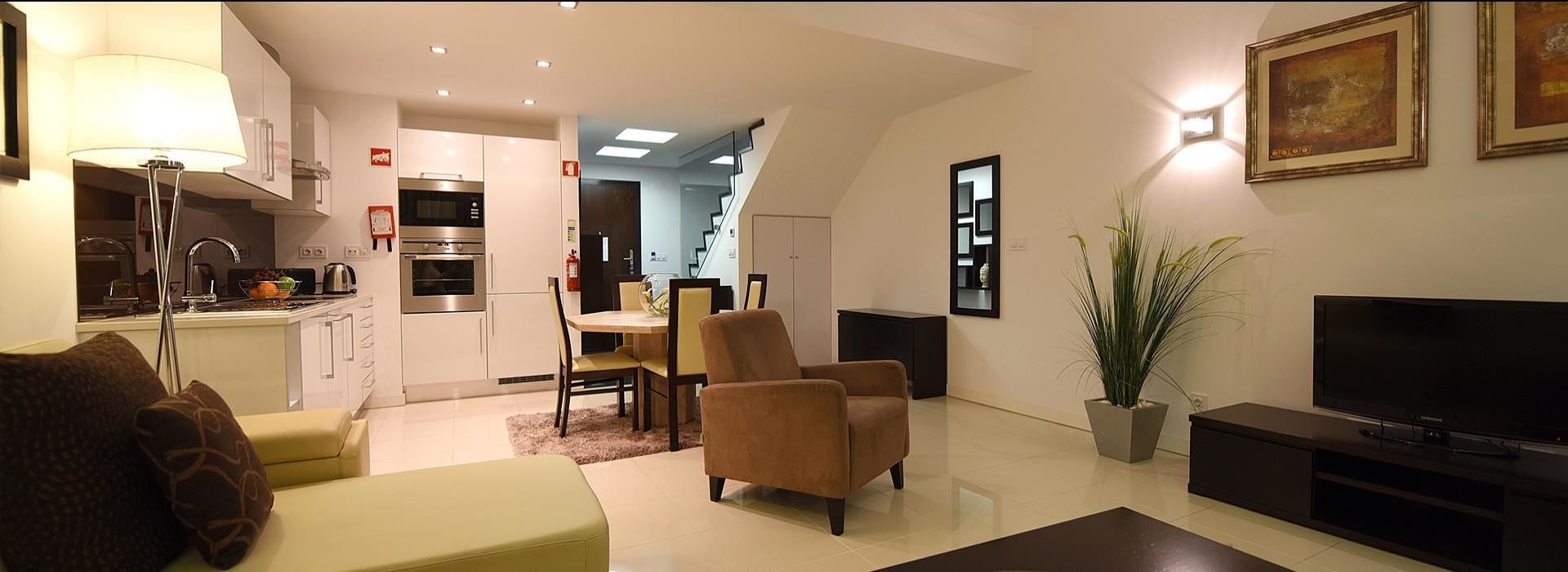 Een moderne woonkamer met een bank, fauteuil en een televisie. Er is een keuken met apparatuur en een eethoek op de achtergrond.