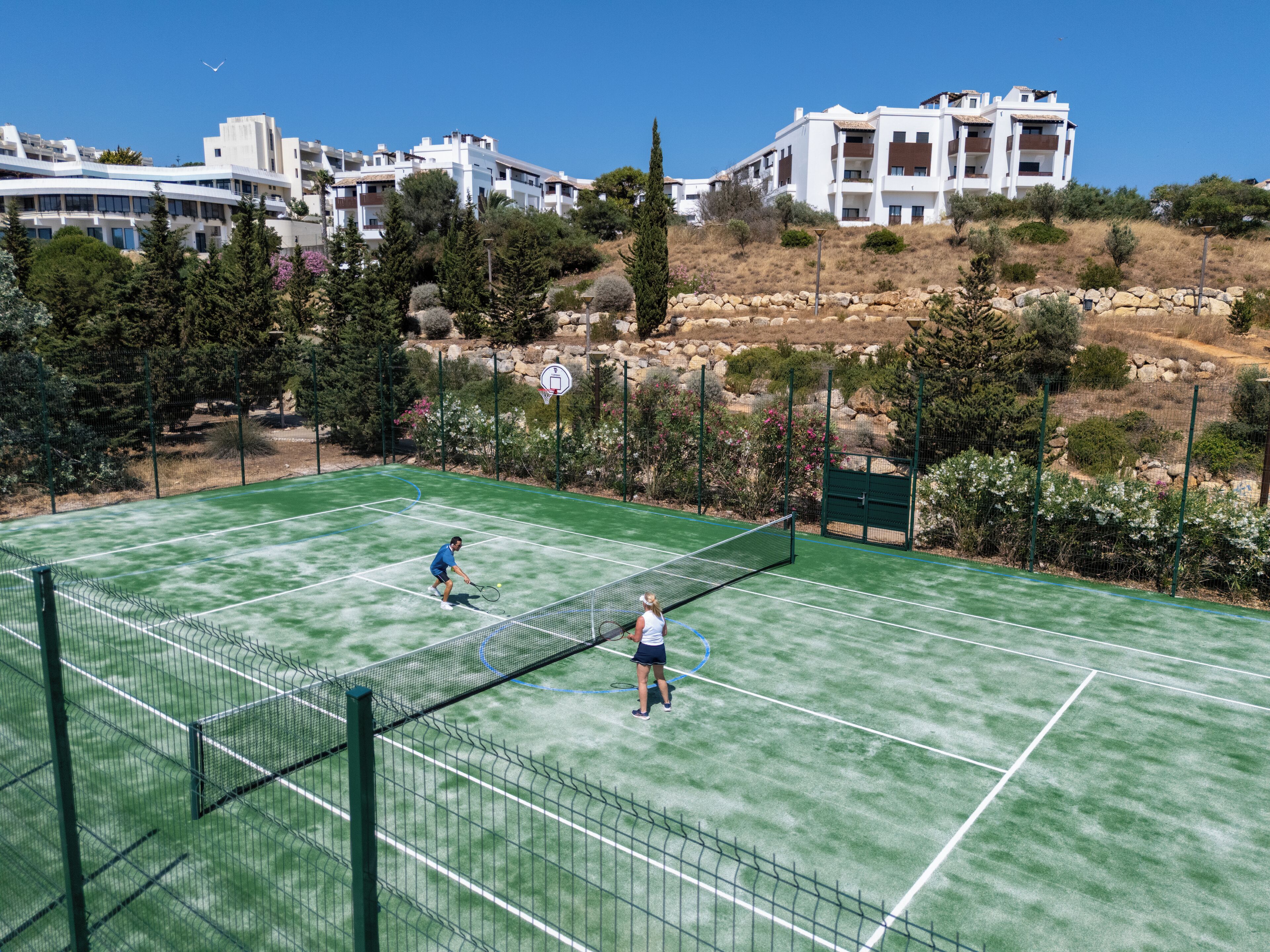 Una cancha de tenis con una red en el centro, rodeada de árboles y edificios al fondo.