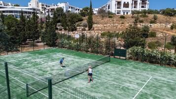 Una cancha de tenis con una red en el centro, rodeada de árboles y edificios al fondo.