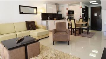 Una sala moderna con un sofá, una mesita y una silla. En el fondo, se ve una cocina con armarios y una mesa de comedor.