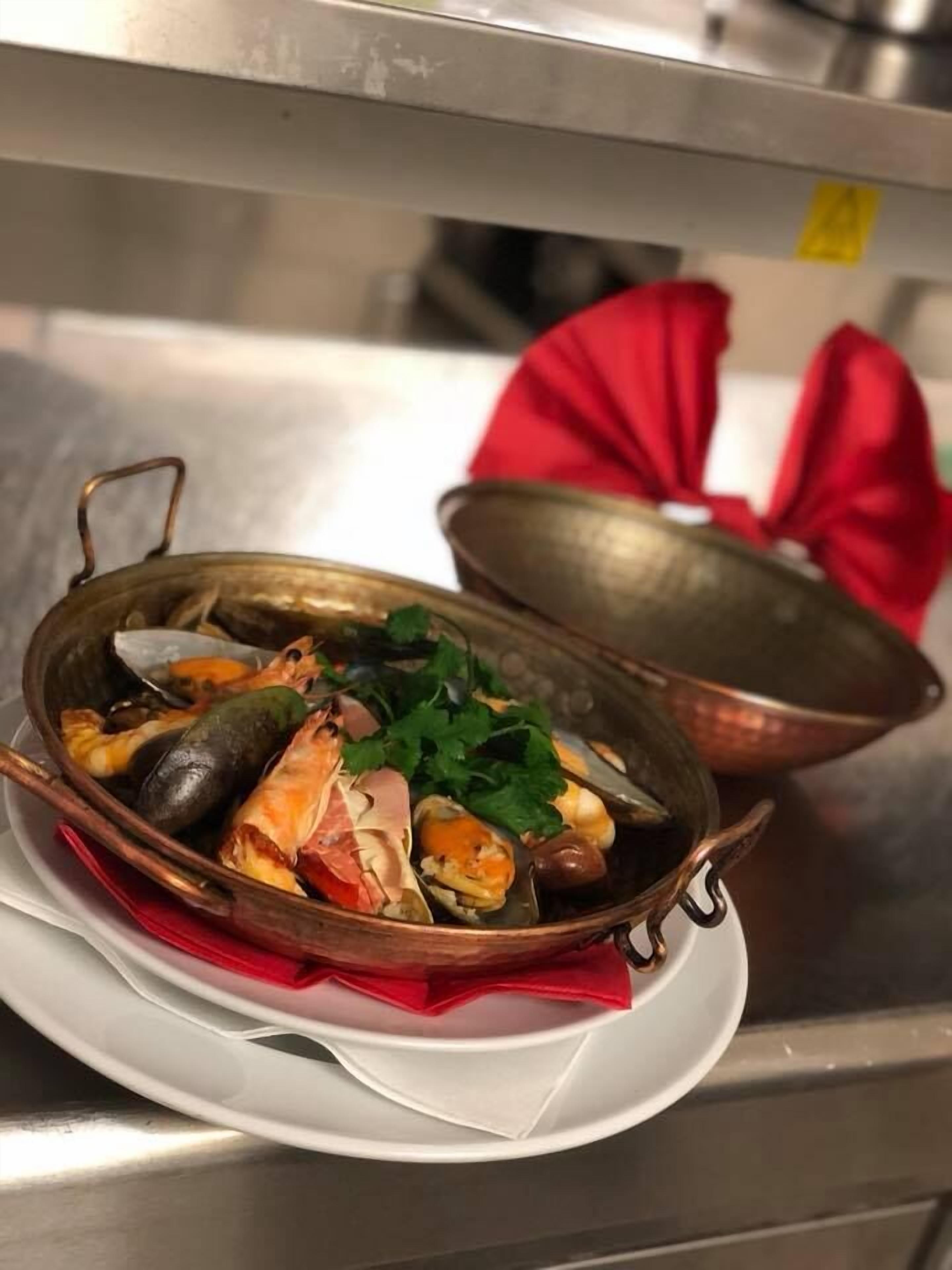 Merellinen ruokalaji tarjoillaan metallisessa astiassa, jossa on kansi. Astia on asetettu valkoiselle lautaselle, ja sen alle on laitettu punainen servetti.