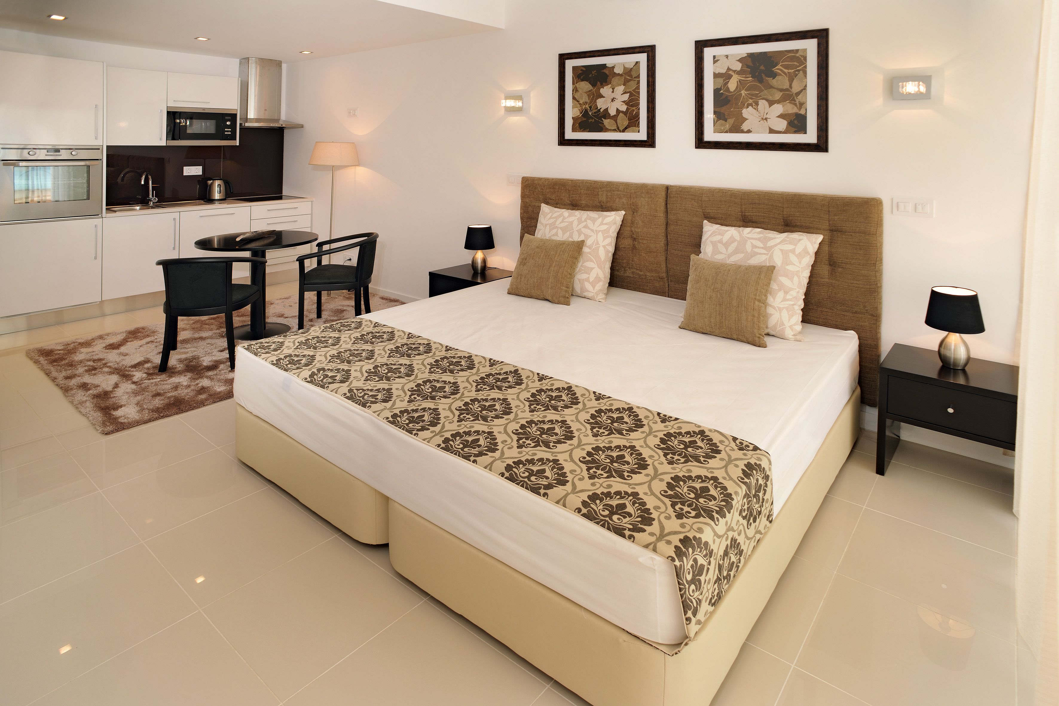 A modern bedroom with a large bed, bedside tables, a desk, and a kitchenette.