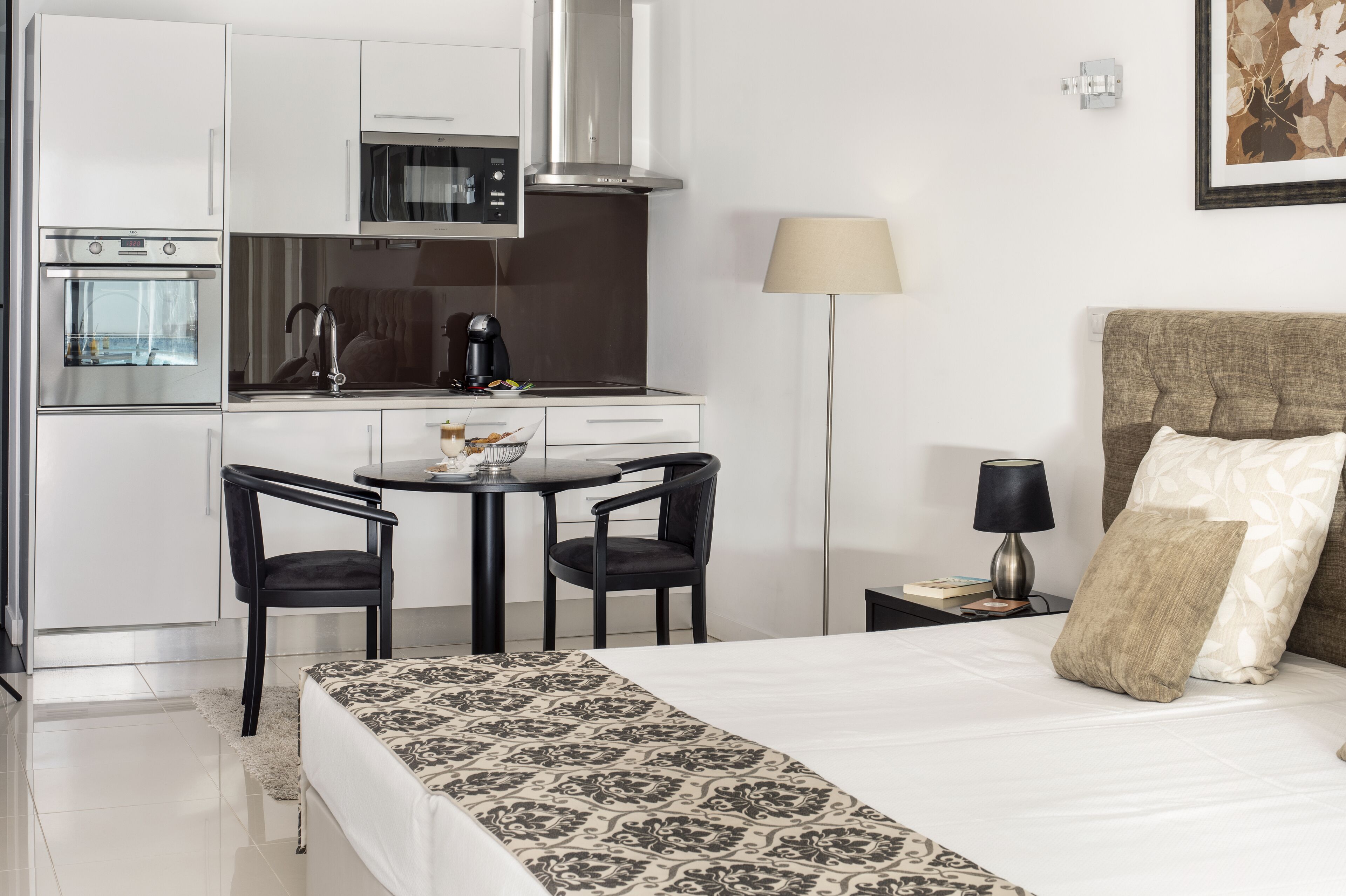 Een compacte hotelkamer met kitchenette, een bed met beddengoed met dessin, een bedlamp en een lijst met bloemistisch kunstwerk aan de muur.