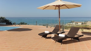 Een terras met twee loungestoelen en een grote parasol, met uitzicht op een heldere blauwe zee en een zwembad.