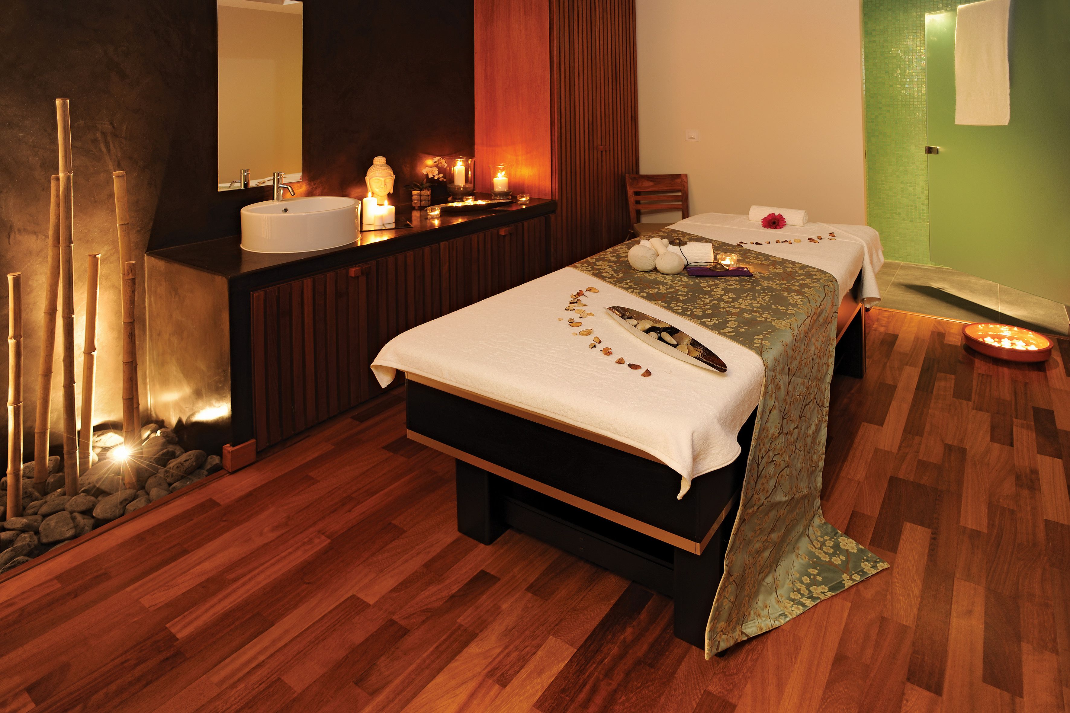 A spa room with a wooden floor, a large bed with a white bedspread, a wooden bench, a sink, and a mirror.