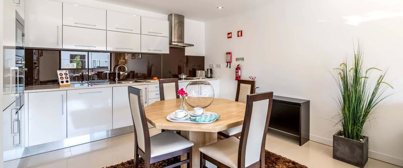 A modern kitchen with white cabinets, a wooden dining table with chairs, and a potted plant.