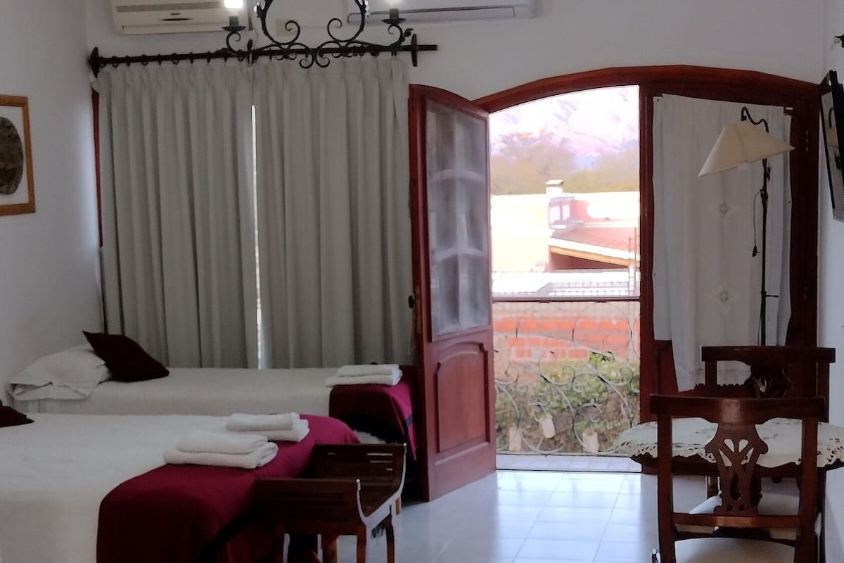 A hotel room with two beds, a chandelier, an air conditioner, a dining table with chairs, and a view of the outside.