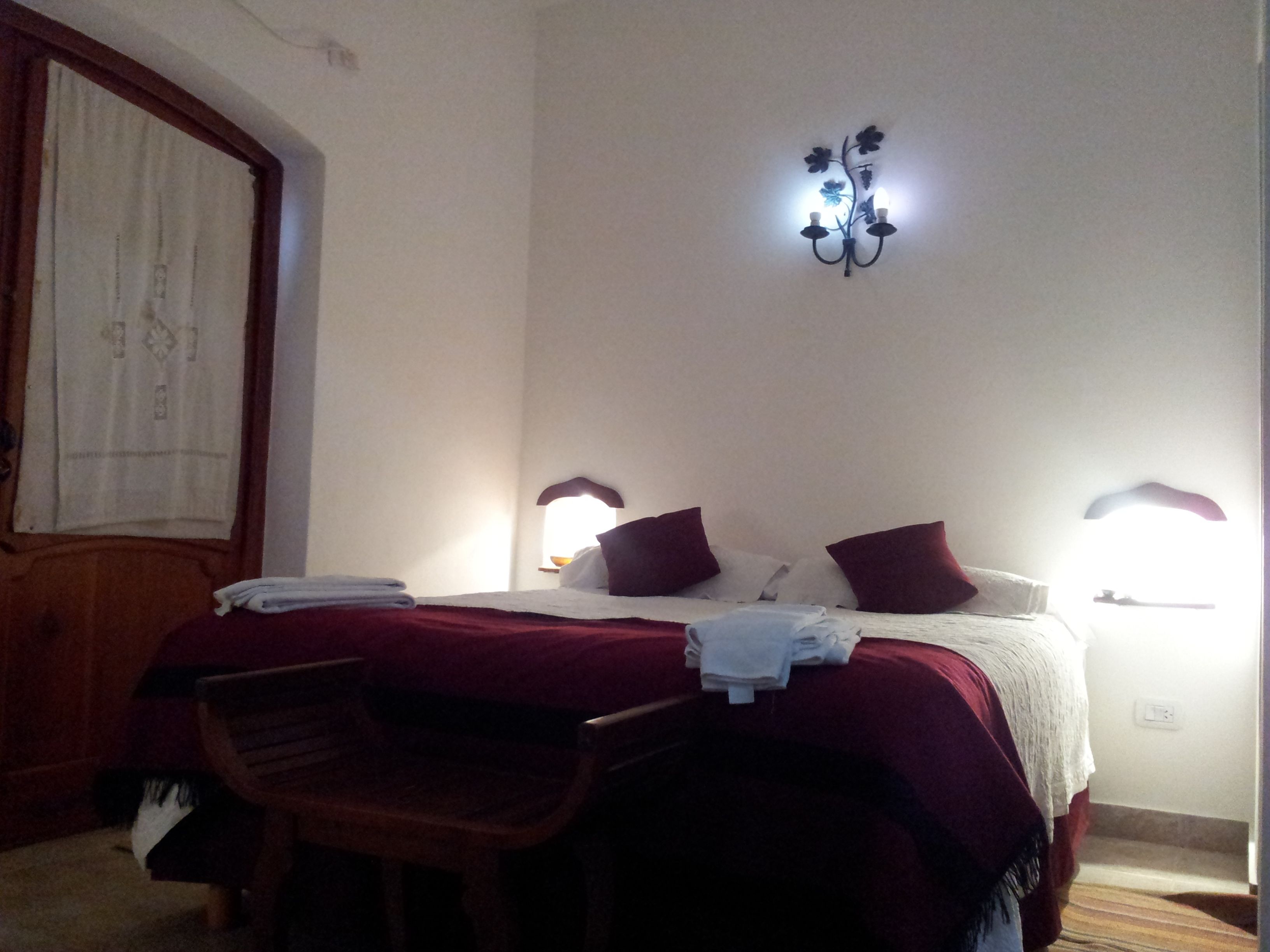 Una habitación de hotel con una cama, dos lámparas de noche, una silla de madera y un aplique de pared.