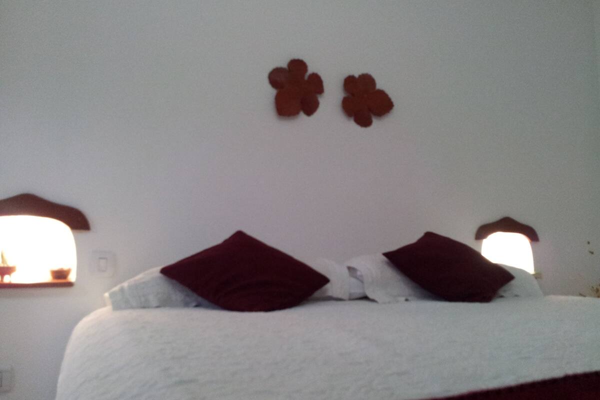 A bed with white bedding and two red pillows, a white wall, and two small windows with brown frames.