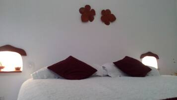 Cama com roupa de cama branca e duas almofadas vermelhas, parede branca e duas janelas pequenas com molduras marrons.