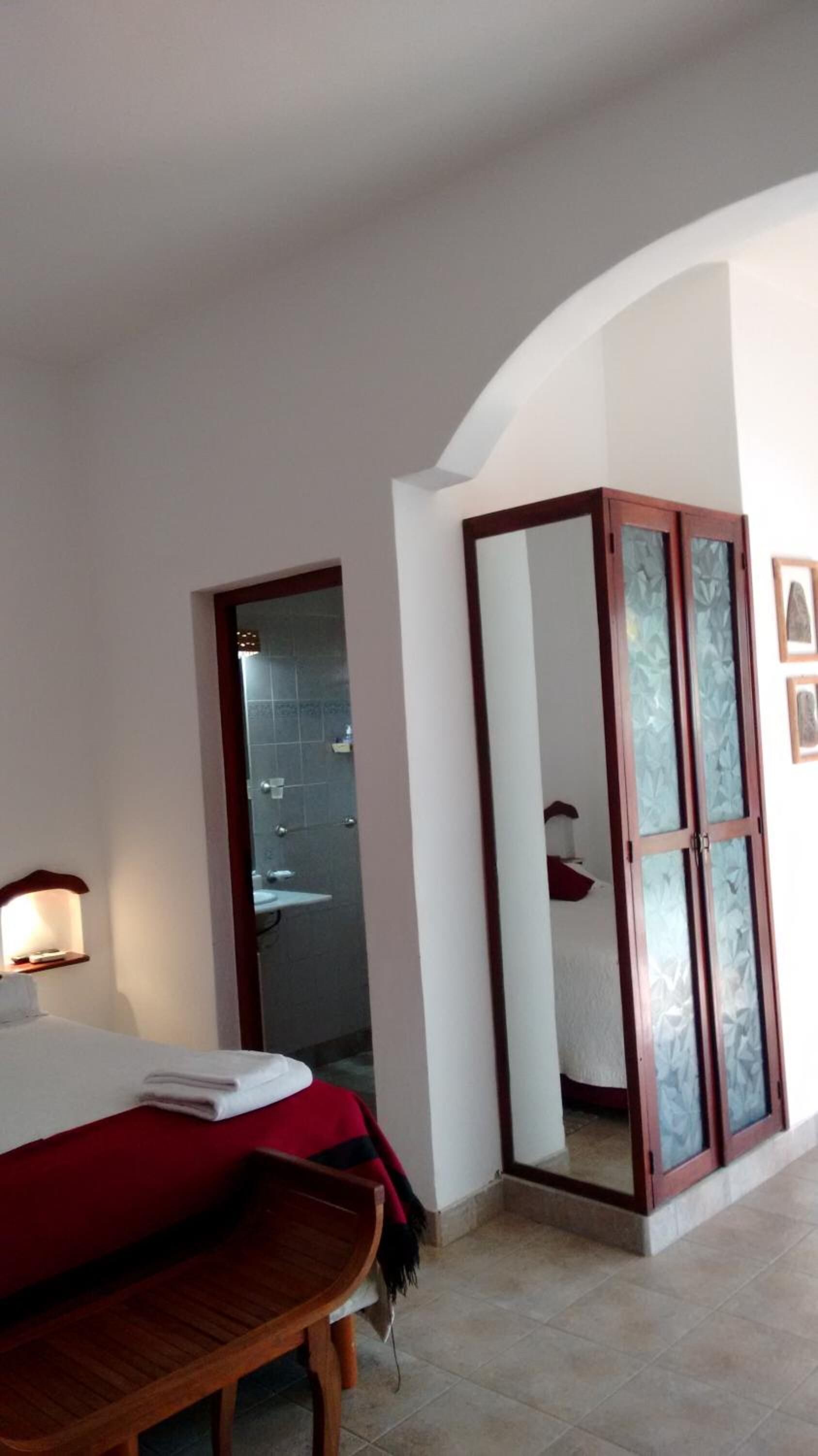 A hotel room with a bed, a wooden chair, a bathroom with a shower, and a glass door leading to another room.